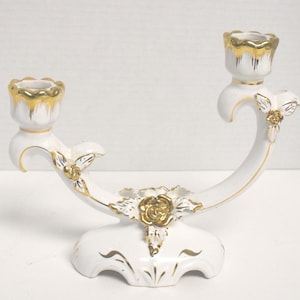 Peut inclure: Un bougeoir double en céramique blanche doté de deux coupelles festonnées aux bords dorés. Les bras incurvés sont ornés de motifs floraux rehaussés d'or, incluant une rose centrale et des feuilles détaillées sur chaque côté.