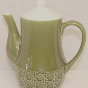 May include: A sage green ceramic teapot featuring a smooth upper body and a textured lower section with a raised woven cane pattern. The teapot includes a curved handle, a tapered spout, and a removable white lid with a pointed finial top.