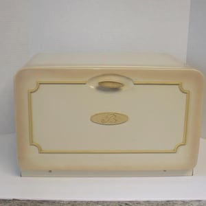 May include: A cream-coloured bread bin with a rectangular shape and rounded corners. The front panel features a gold-toned handle, a decorative gold-coloured border, and a central oval emblem displaying the letter B in a cursive script font.
