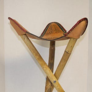 May include: A folding stool with a leather seat and bamboo legs. The tan leather seat features a carved design and red stitching. The bamboo legs are joined with metal hardware, creating a portable seat. A unique, handcrafted item.