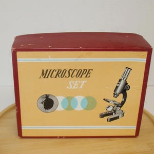 Puede incluir: Una caja de cartón rectangular para un juego de microscopio con un borde granate y un panel central amarillo. El texto MICROSCOPE SET está impreso en blanco y negro. Aparece una ilustración en blanco y negro de un microscopio y un gráfico de insecto.