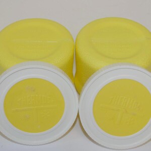 May include: Four yellow Thermos brand lids and two yellow bases. The lids are white with a yellow centre and the word "THERMOS" is printed on the yellow centre. The bases are yellow and have a textured surface.