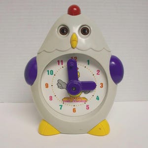 Peut inclure: Une horloge fantaisie en forme de poulet, de couleur gris clair, violet, jaune et rouge. Le cadran de l'horloge présente un design coloré avec le texte "TALKING TIME TEACHER". Les aiguilles de l'horloge sont violettes.