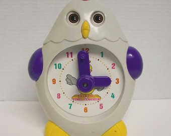 May include: A novelty chicken-shaped clock in light gray, purple, yellow, and red. The clock face has a colorful design with the text "TALKING TIME TEACHER". The clock hands are purple.