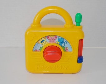May include: A yellow Fisher-Price plastic toy radio with a handle, a green antenna, and a red dial. The center features a colorful illustration of cartoon animals, including a yellow lion, a pink mouse, and a blue mouse, set against a green landscape.