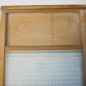 May include: A vintage wooden washboard with a light-coloured wood frame. The top section has faded text, and the lower section features a ribbed, light blue glass surface for washing clothes. A classic laundry tool.