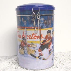 May include: A blue metal tin with a hinged lid, decorated with a hockey scene and the text "Tim Hortons." The illustration shows hockey players and a goalie. The lid is secured with a silver clasp.