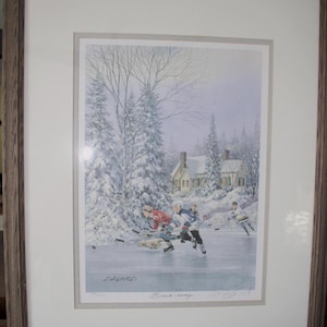 May include: Framed print depicting a winter hockey scene. Players in red and blue jerseys skate on a frozen pond, with a snow-covered landscape and a house in the background. The print is in a brown wooden frame.