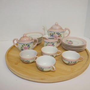 May include: A vintage porcelain tea set with a pink and floral design. The set includes a teapot, sugar bowl, creamer, saucers, and cups. The tea set is arranged on a round wooden tray.