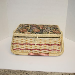 May include: A rectangular sewing basket with a floral patterned lid and woven sides. The lid is padded and trimmed with a cream-coloured fringe. The basket's sides feature a woven pattern with red and cream-coloured stripes. A cream-coloured handle is attached to the sides.