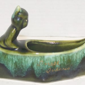 May include: A ceramic canoe-shaped planter in shades of green and brown. A stylized figure is at the back of the canoe. The planter has a glossy finish and the words "Forest Ontario" are written on the side.