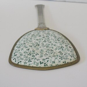 May include: A hand mirror with a triangular shape and a metallic handle. The back of the mirror features a white surface decorated with a dense, repeating pattern of small green flowers, leaves, and vines, enclosed by a thin metallic frame.