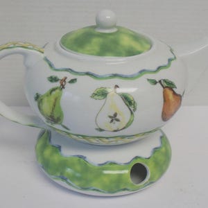 May include: White ceramic teapot with a green lid and base, decorated with pear illustrations. The teapot has a curved handle and spout. The design includes a wavy blue and yellow trim. The teapot sits on a matching warmer.