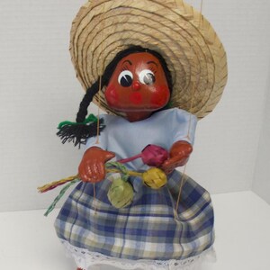 Könnte beinhalten: Handgefertigte Marionettenpuppe mit Strohhut, blauem kariertem Rock und hellblauem Oberteil. Die Puppe hat ein bemaltes Gesicht mit rosigen Wangen und hält einen Strauß bunter Blumen. Die Puppe ist etwa 30 cm groß.