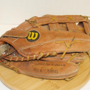 May include: A brown leather softball glove with a Wilson logo featuring a yellow "W" on a black circle. The glove is stitched with tan thread and has the text "Designed for Softball" and "OG 6 A9845" on the palm.