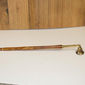 May include: A long-handled candle snuffer featuring a polished brass bell at one end and a smooth, cylindrical wooden handle with a natural grain pattern. The metal arm connecting the handle to the bell is slender and finished in matching brass.