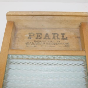 May include: A vintage wooden washboard with a light brown frame and a glass scrubbing surface. The top section features the word "PEARL" and "MANUFACTURED BY CANADIAN WOODENWARE CO." in black text. The glass has a textured, ridged design.