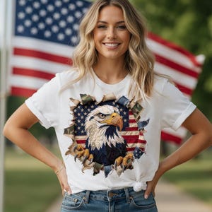 May include: White t-shirt with a patriotic eagle design, incorporating the American flag. The eagle is depicted with a yellow beak and detailed feathers. The shirt is worn with blue jeans. The background includes an American flag.