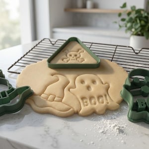 Puede incluir: Tres cortadores de galletas de plástico verde oscuro con formas de sombrero de bruja, un fantasma con el texto BOO y un triángulo con una calavera y tibias cruzadas. Los cortadores están dispuestos alrededor y sobre masa de galleta beige.