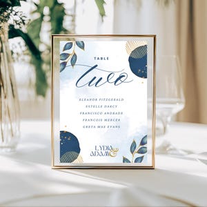 Könnte beinhalten: Eine goldgerahmte Tischkarten. Die Karte zeigt das Wort „Two“ in Schreibschrift, darunter die Namen der Gäste. Marineblaue und goldene Blattmotive verzieren die Karte, mit den Namen des Paares unten. Ideal für Hochzeiten oder Veranstaltungen.