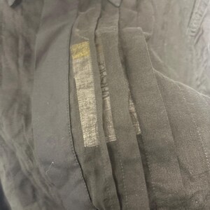 K&ouml;nnte beinhalten: Nahaufnahme eines schwarzen Button-Down-Hemds. Das Hemd hat einen Kragen und eine vertikale Bahn, auf der der Markenname "VERSACE" zu sehen ist. Der Stoff scheint leicht und leicht transparent zu sein.