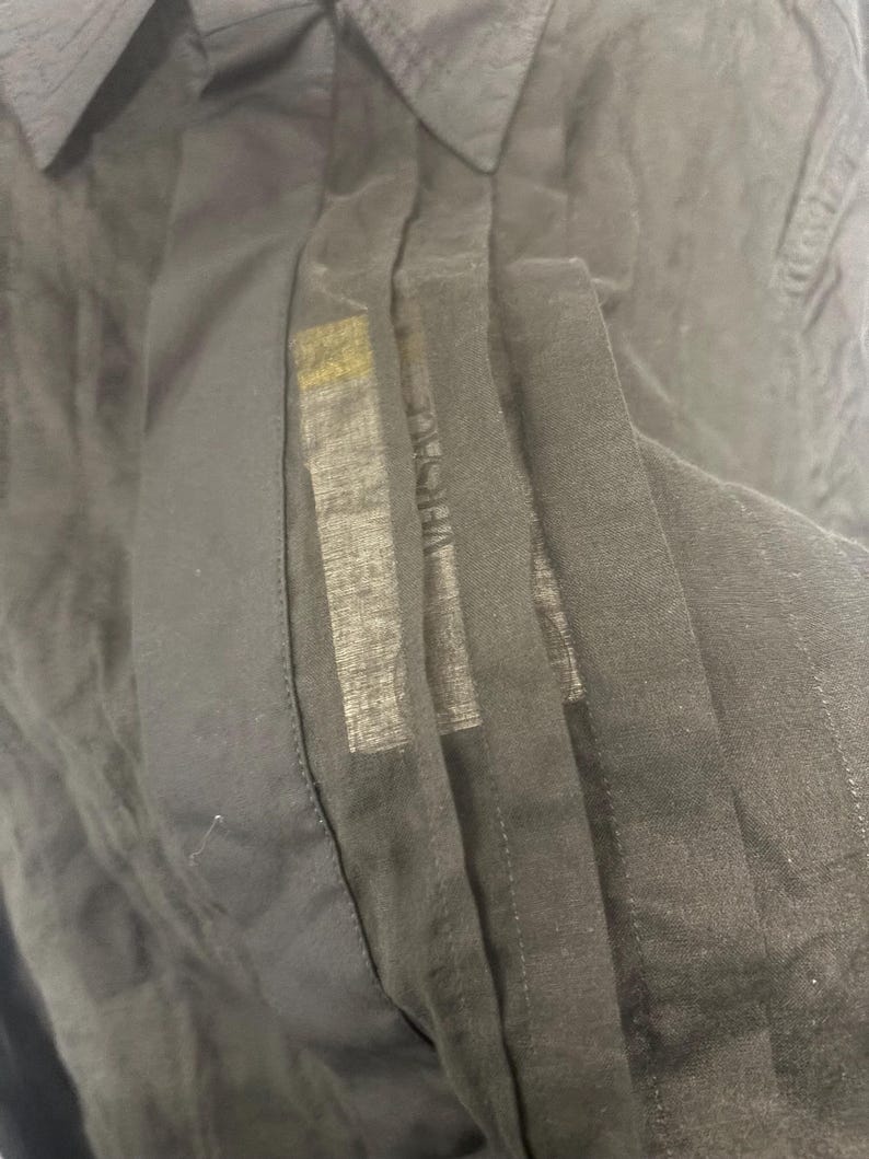 K&ouml;nnte beinhalten: Nahaufnahme eines schwarzen Button-Down-Hemds. Das Hemd hat einen Kragen und eine vertikale Bahn, auf der der Markenname "VERSACE" zu sehen ist. Der Stoff scheint leicht und leicht transparent zu sein.