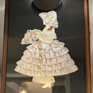 May include: Framed artwork depicting a woman's silhouette in a layered satin dress and hat, adorned with lace and a floral bouquet. The piece is set against a black backdrop, framed in gold.