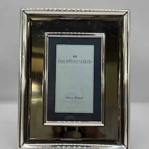 May include: A silver-plated photo frame with a rectangular shape. The frame has a shiny, reflective surface and a dark blue interior. The text "PHILIP WHITNEY LTD." is visible at the top, with "Silver-Plated" and dimensions of 10 cm x 15 cm below.