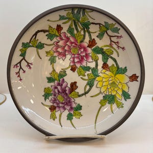 May include: A decorative ceramic bowl with a dark brown rim and a white interior. The bowl features a floral design with pink, purple, and yellow peonies, green leaves, and brown stems. The bowl is supported by a white stand.