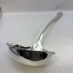 Könnte beinhalten: Eine glänzende, silberfarbene Kelle mit einem langen, gebogenen Griff. Die Kelle hat eine tiefe, abgerundete Schüssel zum Schöpfen von Flüssigkeiten. Der Griff ist glatt und verjüngt sich leicht zum Ende hin. Die Kelle ist aus einem reflektierenden Metall gefertigt.