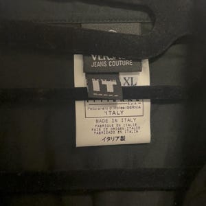 K&ouml;nnte beinhalten: Schwarzes Kleidungsst&uuml;ck mit einem Etikett mit der Aufschrift "VERSACE JEANS COUTURE" und "MADE IN ITALY". Das Etikett zeigt auch die Gr&ouml;&szlig;e "XL" und Text auf Italienisch und Japanisch.