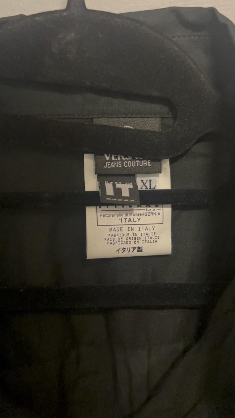 K&ouml;nnte beinhalten: Schwarzes Kleidungsst&uuml;ck mit einem Etikett mit der Aufschrift "VERSACE JEANS COUTURE" und "MADE IN ITALY". Das Etikett zeigt auch die Gr&ouml;&szlig;e "XL" und Text auf Italienisch und Japanisch.