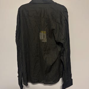 K&ouml;nnte beinhalten: Schwarzes Hemd mit langen &Auml;rmeln, Kragen und gekn&ouml;pften Manschetten. Das Hemd h&auml;ngt an einer wei&szlig;en Wand. Ein kleines, rechteckiges Etikett ist auf der R&uuml;ckseite des Hemdes sichtbar.