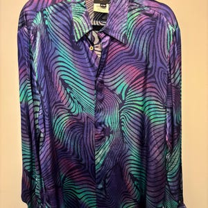 May include: A long-sleeved button-down shirt with a vibrant, swirling pattern in shades of purple, teal, and pink. The shirt has a classic collar and cuffs, and a label is visible at the neck. The fabric has a shiny, silky appearance.