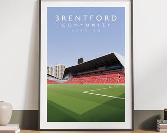 May include: Framed print of the Brentford Community Stadium, showing a green pitch, red seating, and the stadium's name in white text against a blue sky. The artwork is displayed on a wooden surface.