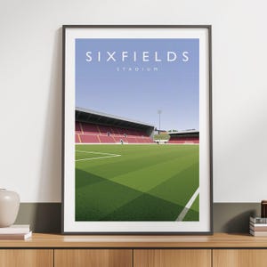 Peut inclure: Impression encadrée du stade Sixfields, avec un terrain vert et des lignes blanches, des sièges rouges et un ciel bleu. Le nom du stade est en blanc. Un lieu sportif moderne.