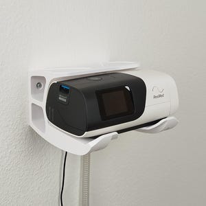 Op de afbeelding: Een wit en zwart CPAP-apparaat met een digitaal display, gemonteerd op een witte wandbeugel. Het apparaat is voorzien van het label "ResMed" en is aangesloten op een slang. De beugel is bevestigd aan een witte muur.