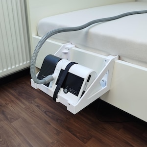 Może przedstawiać: Biały uchwyt na maszynę CPAP przy łóżku przymocowany do ramy łóżka. Uchwyt podtrzymuje czarno-białą maszynę CPAP z podłączonym szarym wężem. Uchwyt jest wykonany z białego drewna i zamontowany na drewnianej podłodze.