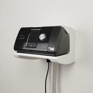 Op de afbeelding: Een zwarte AirSense 10 Auto CPAP-machine gemonteerd op een witte wandplank. De machine heeft een digitaal display, een zilveren bedieningsknop en het ResMed-logo. Een zwarte slang en een witte geribbelde buis lopen naar beneden.
