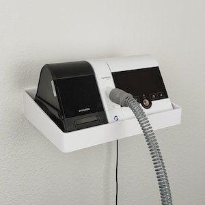 Puede incluir: Un dispositivo médico blanco y negro, montado en un estante blanco, contra una pared blanca. El dispositivo tiene una manguera gris conectada. El dispositivo está etiquetado como "primaAQUA" y "LOWENSTEIN".