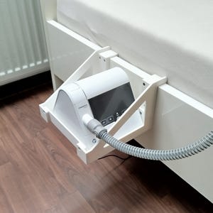 Op de afbeelding: Een wit CPAP-apparaat met een zwart scherm is gemonteerd op een witte beugel die aan een bedframe is bevestigd. Een grijze slang loopt van het apparaat. Het bed heeft een wit laken en staat boven een houten vloer.