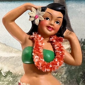 May include: A vintage figurine of a woman in a green top and a red and white lei. She has a flower in her hair and is smiling. The figurine is painted with bright colors and has a tropical theme.