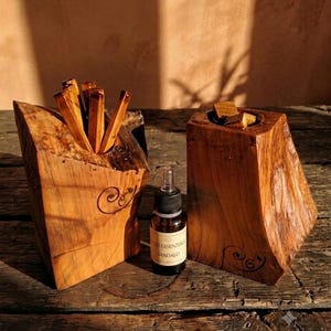 Può includere: Due blocchi di legno naturale con bordi grezzi e organici e venature scure, ciascuno con un'apertura intagliata sulla parte superiore. Un blocco contiene diversi bastoncini di legno sottili, l'altro piccoli pezzi di legno. Un flacone contagocce in vetro con etichetta OLIO ESSENZIALE SANDALO si trova tra i due.