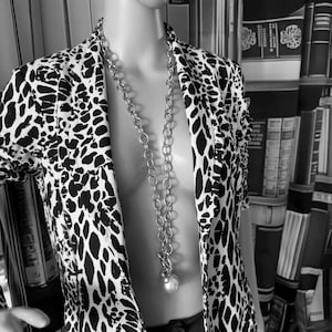 Könnte beinhalten: Ein schwarz-weiß gemusterter Blazer mit Tiermuster über einem einfarbigen schwarzen Kleidungsstück, dazu eine lange silberfarbene Gliederkette mit großen runden Kettengliedern und einem weißen perlenartigen Anhänger an einem Knebelverschluss.