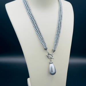 Può includere: Una collana a tre fili composta da piccole perle rotonde color grigio argento metallizzato. I fili sono collegati da una chiusura a T argentata, che sostiene un unico grande ciondolo a forma di goccia con una finitura grigio argento.