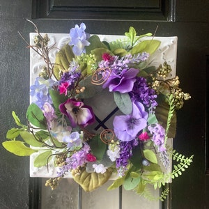 May include: A purple, white, and green floral wreath with lavender accents. The wreath is made of silk flowers and is arranged on a white background.