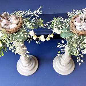 Pu&ograve; includere: Due portacandele in legno grigio con un nido di uccelli e uova in cima a ciascuno. I nidi sono circondati da ramoscelli verdi e bianchi.