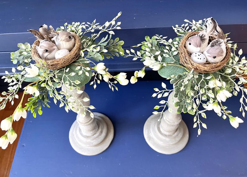 Pu&ograve; includere: Due portacandele in legno grigio con un nido di uccelli e uova in cima a ciascuno. I nidi sono circondati da ramoscelli verdi e bianchi.