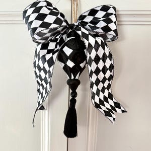 May include: A black and white checkered bow with long ribbon tails, a black teardrop-shaped ornament, and a black tassel. The bow is hanging from a natural twine loop, suitable for home decor.