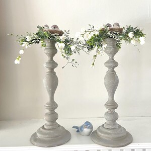 Pu&ograve; includere: Due portacandele in legno grigio con una ghirlanda floreale bianca e verde e piccoli nidi di uccelli in cima. I portacandele sono su uno scaffale bianco.